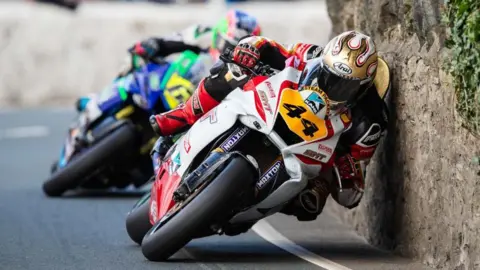 SOUTHERN 100 Riders leaning into a stone wall on a bend, the bikes and leathers are colourful and the bike at the front at the number 44 in black on a yellow plate on the front.