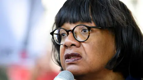 Diane Abbott, who has medium length dark hair and round glasses, speaking into a microphone.
