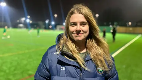 Elizabeth Baines / BBC A woman with blonde hair stands on a football pitch. She wears a blue puffer coat