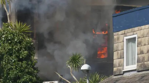Naked flames behind a window of the hotel. There is thick black smoke inside and outside of the former hotel.