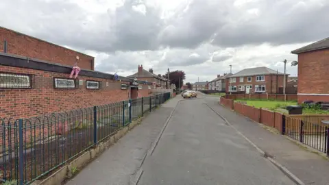 Google Millbrook Avenue is a residential road. A red-bricked social club is on the left, with a blue metal fence in front of it. Two-storey red bricked semi-detached houses line the road further down.