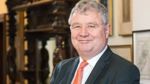Isle of Man Government Chris Thomas has short grey hair. He's wearing a dark suit jacket, white shirt and orange tie. He's standing in a room with pictures handing on the wall to the right, and a dark wooden piece of ornate furniture on the left.