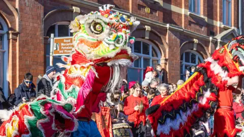 Museum of Royal Worcester Large colourful costumes tower above crowds watching by a building, which has arched windows. A brown sign includes the words Royal Worcester Porcelain and Museum.