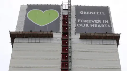 PA Media The top of the Grenfell tower block wrapped in a covering with a large green heart and a sign that reads Grenfell Forever in our Hearts