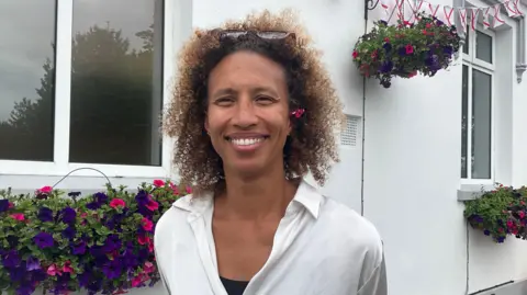 A woman with curly brown hair, wearing a white blouse. She has a pair of sunglasses on her head and a flower tucked behind her left ear. She is staring directly at the camera and smiling. Behind her are a number of hanging baskets with flowers in them, plus bunting with Jersey flags.
