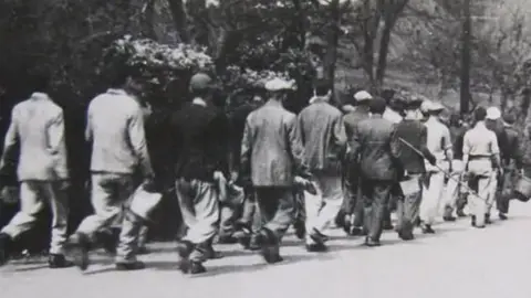 Alderney Museum Black and white image of men dressed in dirty clothes and boots being marched along the road