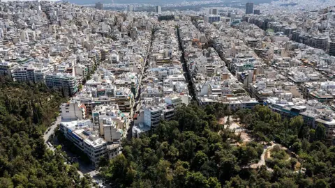 Aerial view of a densely built urban area with rows of white and beige apartment buildings stretching across the landscape. In the foreground, a large green park filled with mature trees contrasts with the surrounding city blocks.