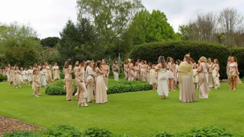 Mark Johnston A wide shot of the bridesmaids standing in a grassy area. They are all wearing gold dresses.