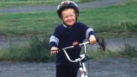 Selena Bostock. Una foto borrosa muestra a un jovencísimo Matthew vestido de azul oscuro, sosteniendo una bicicleta plateada y con casco. Está de pie entre asfalto y césped.
