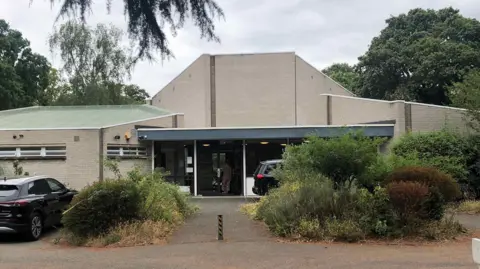 A low, cream-brick building with a covered glass entrance, set back behind shrubs and a gravel drive.