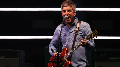 Reuters Noel Gallagher in blue denim sings into a microphone and plays a red guitar at the Oasis comeback gig in Cardiff