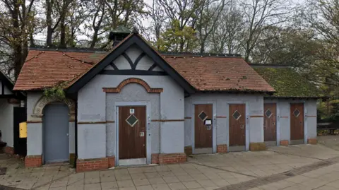 A grey one-storey building is in the middle. It has multiple brown wooden doors and a red roof. There are trees behind the building.