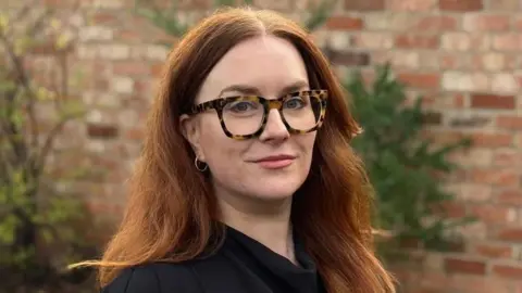 Matilda Robinson-Murphy has long auburn hair. She is standing in front of a brick wall, wearing a black top and glasses with tortoise shell rims.