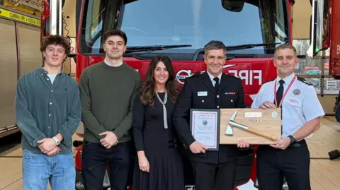 Cumbria Fire and Rescue Service L-R Ashton Hook, Ellis Hook, Jennifer Hook, Stuart Hook and Tommy Seel. Tommy is presenting Stuart with a commemorative axe in front of a fire engine in a control room.