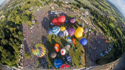 Paul Box The site of the Bristol Balloon Fiesta is seen from the basket of a hot air balloon that has just taken off. Thousands of spectators can be seen on the ground, along with dozens of other balloons which are inflated ready to take off. Fields and the boundary of the city of Bristol can also be seen in the distance