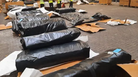 National Crime Agency A haul of rolled up inflatable black boats in a warehouse. Flattened cardboard boxes are littered around them 
