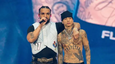 Getty Images They are standing stage with Drakes arm around Central Cee. Drake is speaking into a microphone and has a grey puffer vest on. Central Cee has his top off with his tattoo's showing.