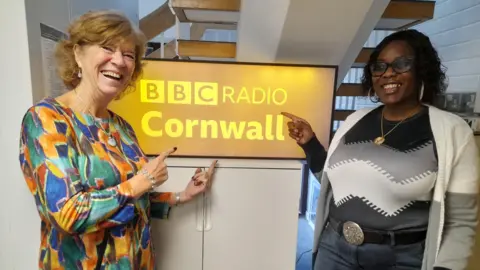 CASS+ Two women smiling whilst pointing at a BBC Radio Cornwall lit up box which is on top of a cupboard and underneath a staircase.