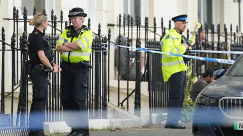 A picture of the crime scene in Percival Terrace, near Marine Parade, in Brighton, East Sussex. Two police officers wearing high-vis can be seen in the image alongside another officer in uniform. A police cordon is also visible.