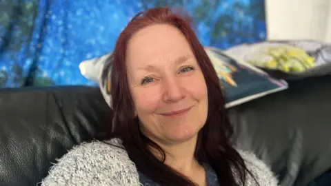 Amber Richards smiles to camera, she has red hair and is sitting on a black sofa with cushions behind her head