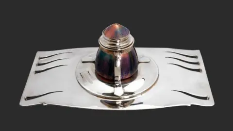 The inkwell, which has a decorative silver base with a colourful pot that has a lid in the middle of it.