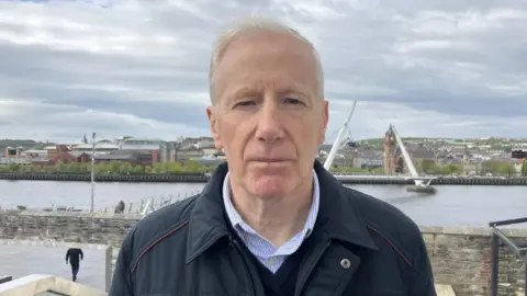 Shows a man in a dark jacket, blue and white striped shirt and blue v-neck jumper standing in front of the Foyle Bridge
