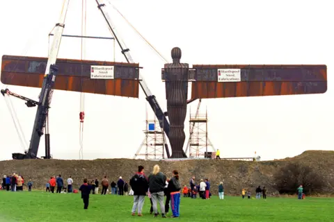 Owen Humphreys / PA Media The wings of the Angel of the North are being hoisted onto its body by black and white metal cranes. Scaffolding is erected on the hillside where the sculpture stands. The 65ft (22m) tall, brown sculpture appears to be a man with his wings outstretched. There are dozens of people standing in the field below looking up at the scene.