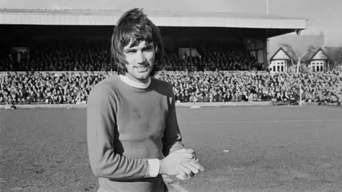 Getty Images George Best facing the camera at a Manchester United match against Northampton in 1970. The black-and-white image shows him on the pitch with a full stand of supporters behind him. 