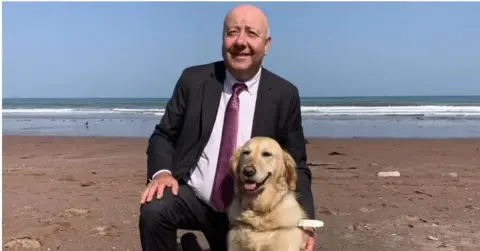 Steve wearing a suit and tie on a beach with a golden labrador Jennie
