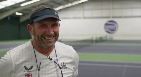 Neil Bamford is standing on the tennis courts and he is staring directly at the camera and laughing. He is wearing a black visor and white tennis T-shirt. 