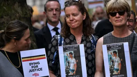 Families protesting outside the infected blood public inquiry in July 2023. Three women are holding pictures of their brother as a young man who died of Aids because of the scandal. 