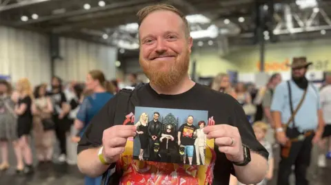 A man is wearing a black top with an orange and red image of Black Sabbath. He is smiling and holding a photograph of him standing with two men and two women.