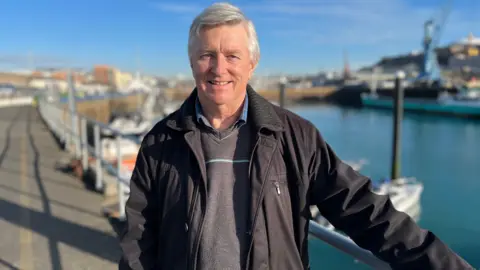 Ian Cuming - a man wearing a grey knit jumper under a black coat standing at the harbour.