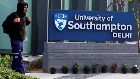 A man wearing a brown hoodie and blue trousers passes by the entrance to the campus. On his side is a board which reads University of Southampton Delhi. There are trees and shrubs on the side and a glass building in the backdrop.