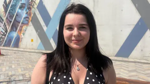 Lauren, She has long dark hair, wearing a black and white polka dot sleeveless top and silver necklace.