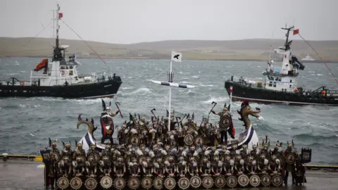 EPA Members of the Up Helly Aa festival's 'Jarl Squad' pose with the replica Viking longship after the morning parade in Lerwick, Shetland Islands. There are also two fishing boats on the picture.