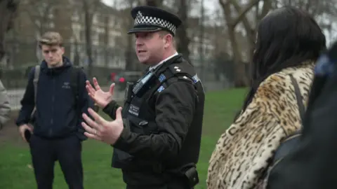 Metropolitan Police Inspector Ollie Lamb is stood in a park wearing his Met Police uniform. He is gesticulating with his hands, and either side of him is a woman and a man. In the background are the black railings of the park, road and buildings.