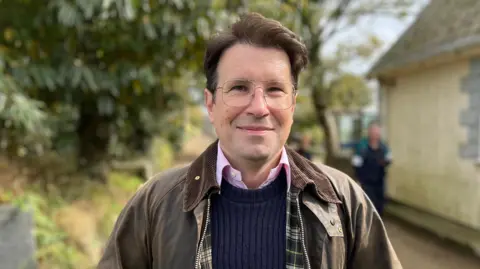 A man with a brown jacket and blue jumper wearing glasses and smiling looking at the camera. 