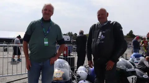 Neil standing with his hand in his pocket in front of his bikes, with another man standing to his left. 