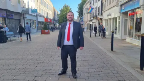 A man in a dark suit and red tie stands in a pedestrianised shopping street. He has short hair and a beard.
