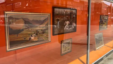 Amy Holmes/BBC A window display on a high street featuring pictures from the Bedfordshire Art Loan collection, which are mounted on an orange background.