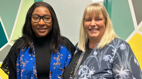 Two teachers stand smiling. The teacher on the left has dark hair and skin. She wears a blue satin short sleeved cardigan with pink flowers and black lace. The teacher on the right is light skinned and has light blonde hair. She wears a grey top with white flowers and reeds on.