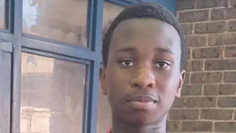 Eghosa stands in front of a blue door and a brick wall, he is sticking his thumbs up and he is wearing a medal around his neck 