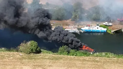 A photo taken from above by a drone shows thick black smoke billowing from a small burning boat in a dark river