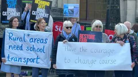 BBC/Claudia Robinson A group of people holding signs which read "rural areas need school transport" and "have the courage to change course" as well as "Settle says bring transport back" and "Richmond says bring catchment back".