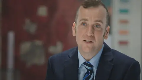 Simon O'Donovan. He has short brown hair, blue eyes, wearing a navy blazer, light blue shirt and blue striped tie.