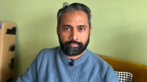 A man, Sree Kadalyil, looks into the camera. He is sitting on an orange sofa that is up against a light green wall. He has short, greying black hair and a black beard that is speckled with grey. He is wearing a light blue jumper.