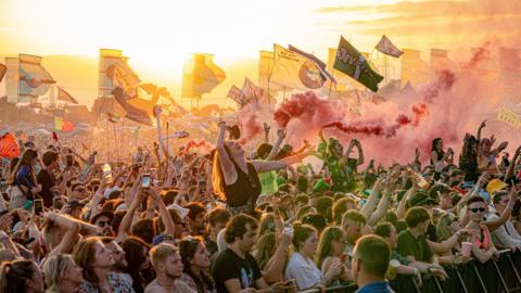 Wide view of the crowd at Glastonbury near sunset, with lots of flags and flare smoke visible. 