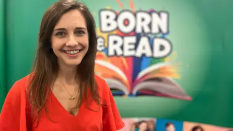 Jon Wright/BBC A woman in a red top stands in front of a sign which reads: Born and Read