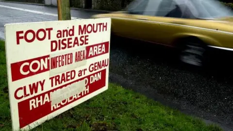 PA Media A sign at the Menai Bridge leading onto Anglesey during the 2001 outbreak warning of "foot and mouth disease infected area". A blurred gold-coloured car drives past it. 